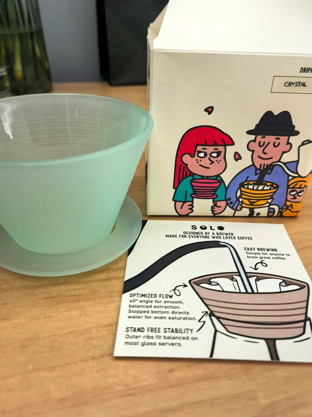 A pale mint-green ribbed coffee dripper sits on a matching saucer beside its illustrated box and an English-language instruction card on a wooden table. The card diagrams water flowing through the dripper and describes its brewing features.