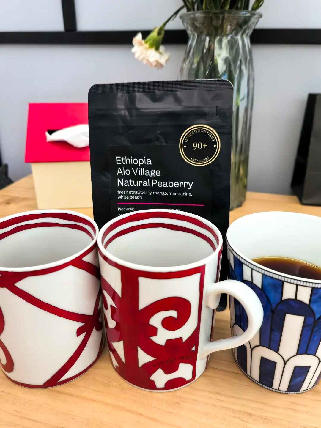 A black coffee bag labeled “Ethiopia Alo Village Natural Peaberry” stands behind three patterned mugs on a wooden table; the right mug contains coffee. A vase of flowers and a tissue box are in the background.