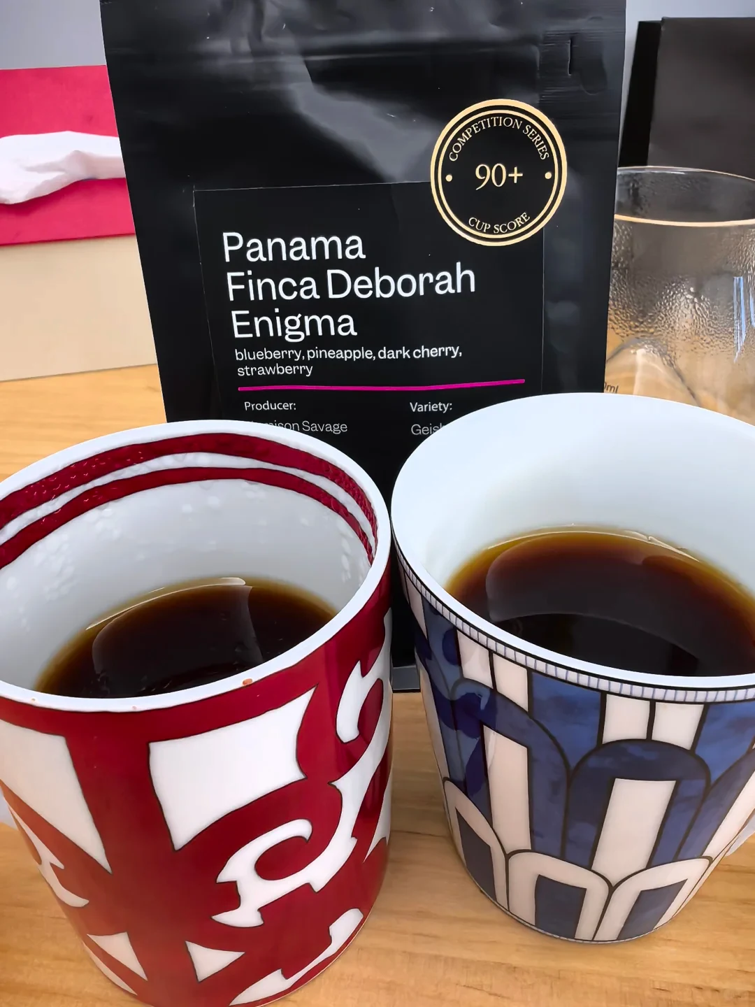 Two patterned ceramic cups of dark coffee sit on a wooden counter in front of a black coffee bag labeled “Panama Finca Deborah Enigma.”