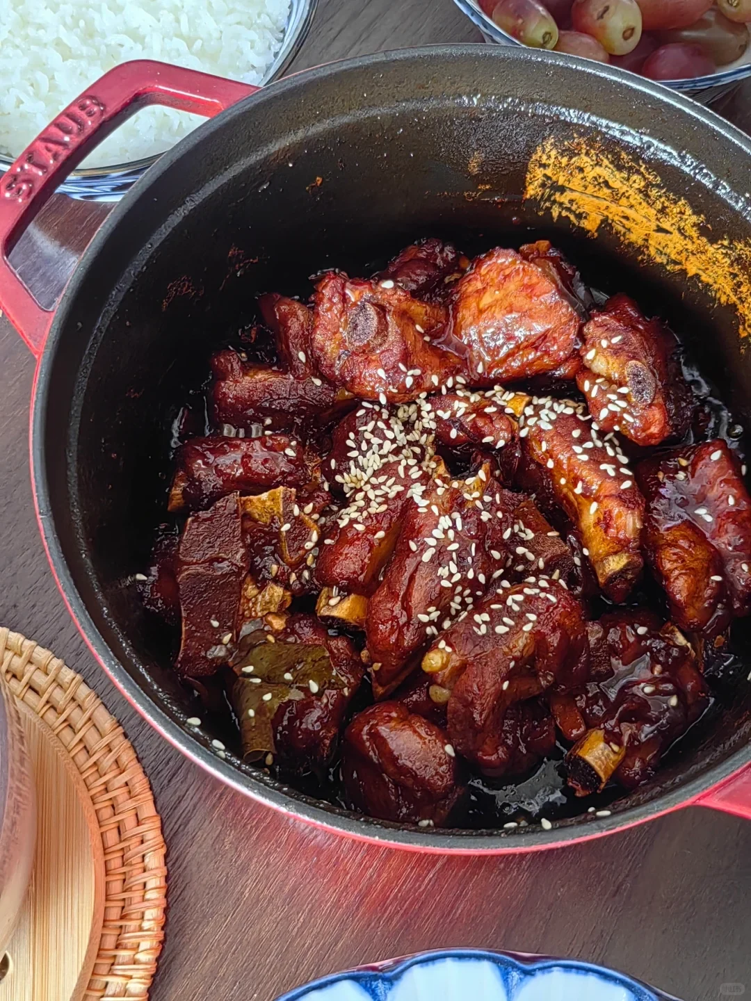 Sweet-and-sour pork ribs sprinkled with sesame seeds in the owner’s red Staub round cocotte, with rice and fruit nearby.