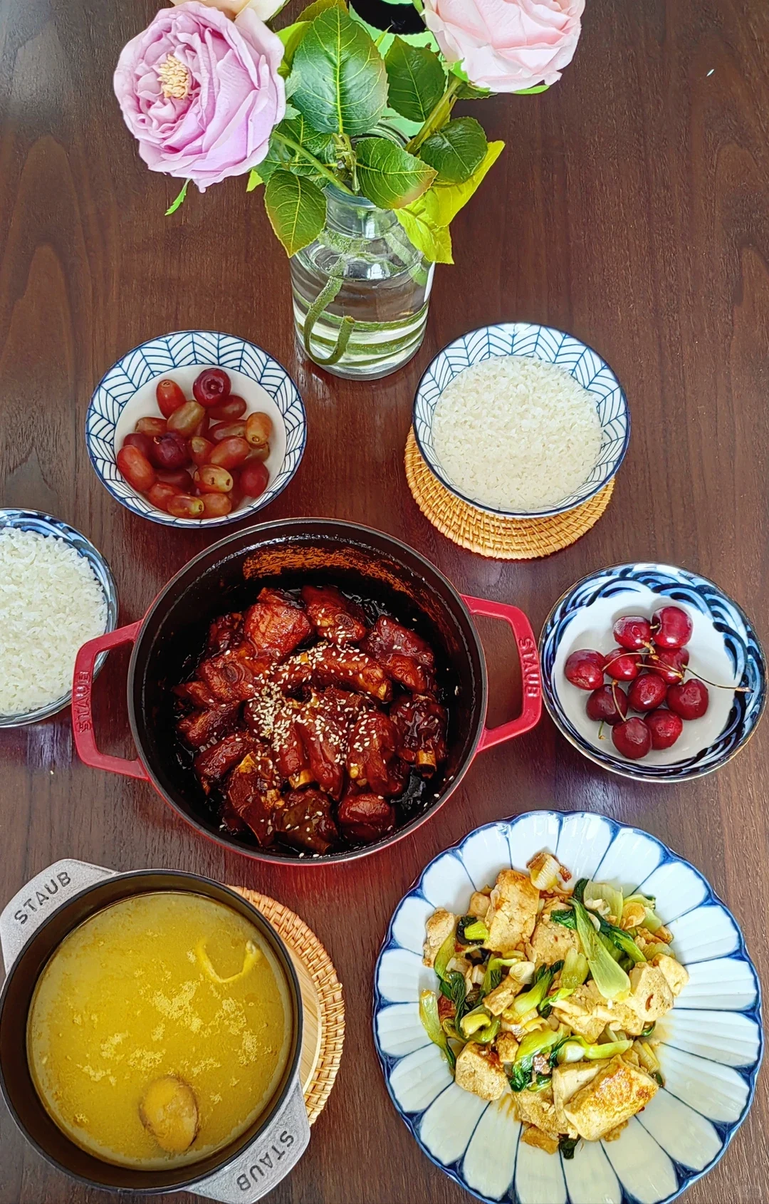 An overhead view of a meal with the red Staub cocotte of pork ribs, rice, fruit, a smaller pale pot of soup, a plate of cooked food and pink flowers.