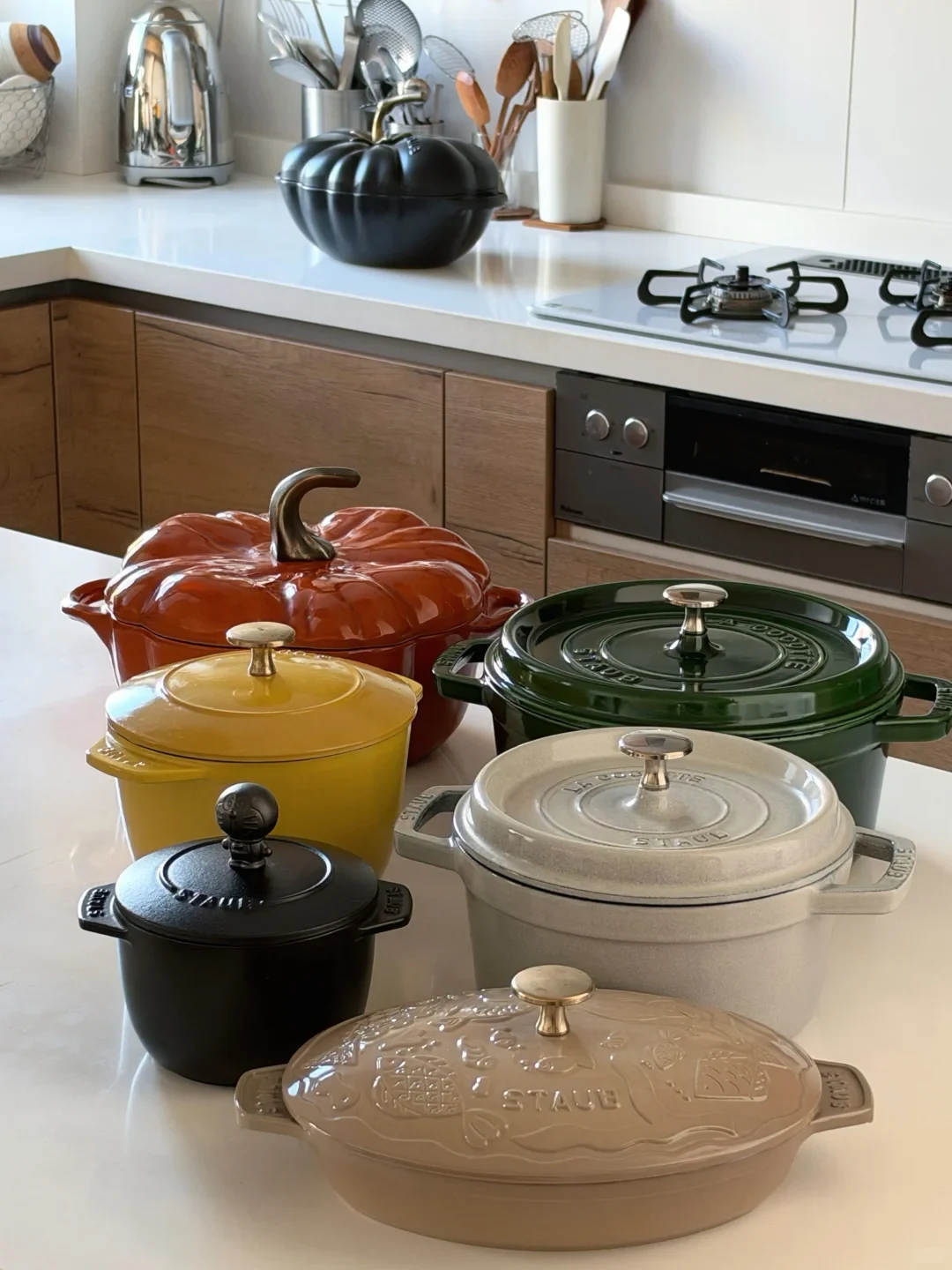A collection of colorful Staub cast-iron cookware arranged on a white kitchen island, including an orange pumpkin-shaped cocotte, a deep green Dutch oven, a yellow pot, an off-white round cocotte, a small black cocotte, and an embossed taupe oval dish, with a modern kitchen and stovetop in the background.