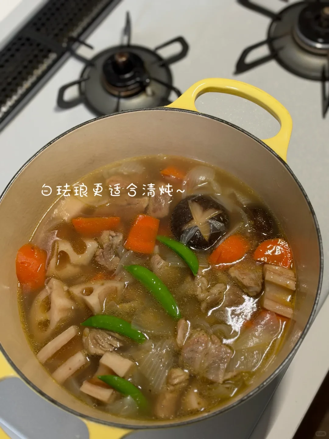 A simmering clear broth stew with lotus root, carrots, shiitake mushrooms, green beans, and meat cooked inside a yellow enameled pot on a stovetop.