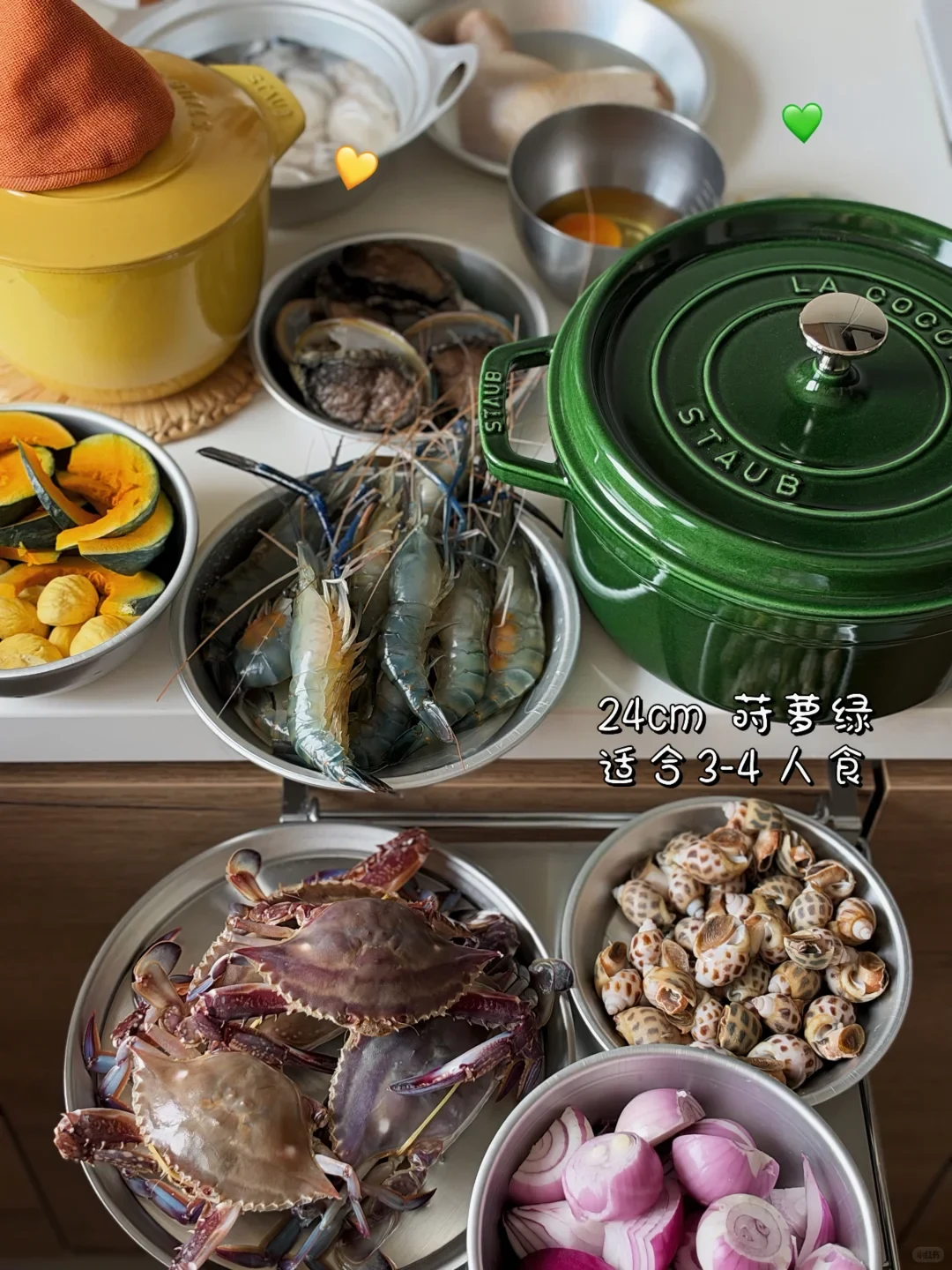 Ingredients for a seafood feast prepared in metal bowls around a green Staub cast-iron pot, including live river prawns, swimming crabs, sea snails, sliced shallots, pumpkin, and abalone.
