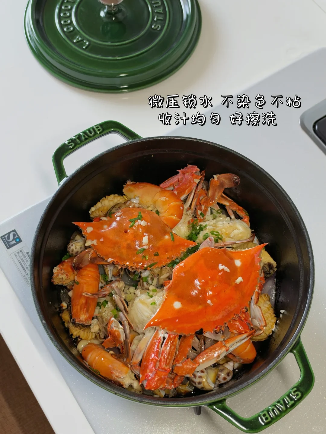 Cooked seafood featuring crabs, shrimp, and shellfish prepared in a green Staub cast iron pot on a stovetop with its matching lid placed nearby.