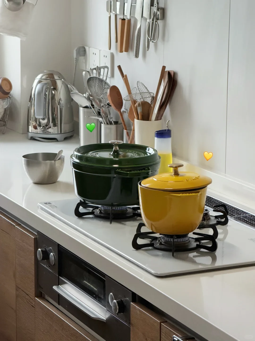 A dark green enameled cast iron Dutch oven and a small yellow cast iron pot on a white gas stovetop with lit burners in a modern kitchen.