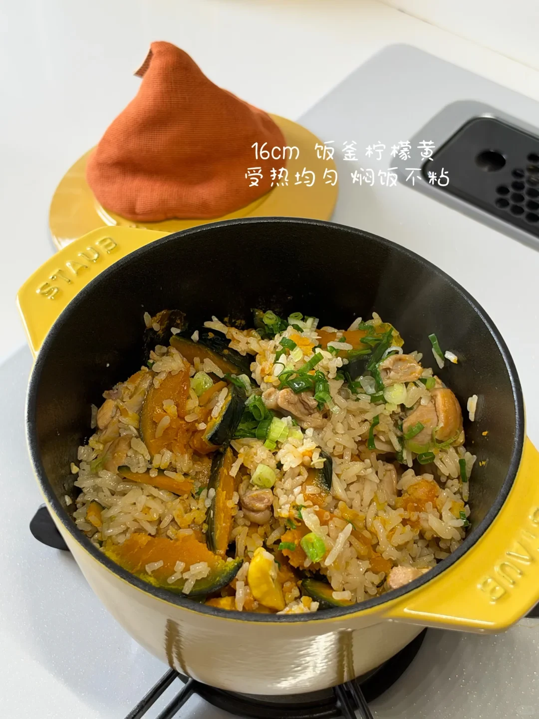 A yellow Staub cast iron rice cocotte filled with cooked pumpkin and chicken mixed rice topped with chopped scallions, resting on a gas stove top.