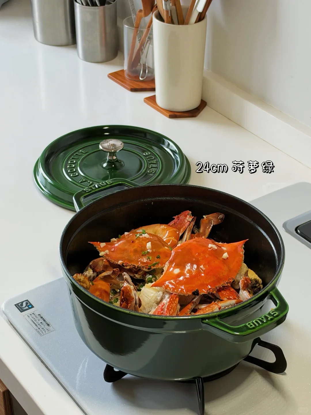 A green Staub cast-iron pot filled with cooked whole crabs sits on a stovetop with its matching lid placed behind it.