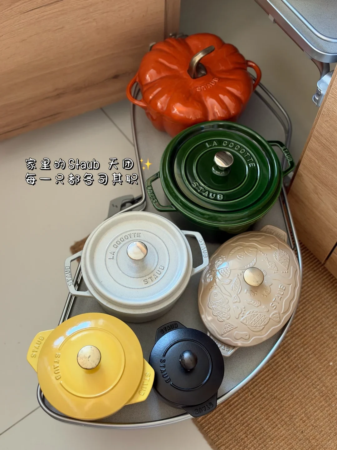 An overhead view of six Staub enameled cast iron pots in various colors, shapes, and sizes arranged inside a pull-out kitchen corner cabinet shelf.