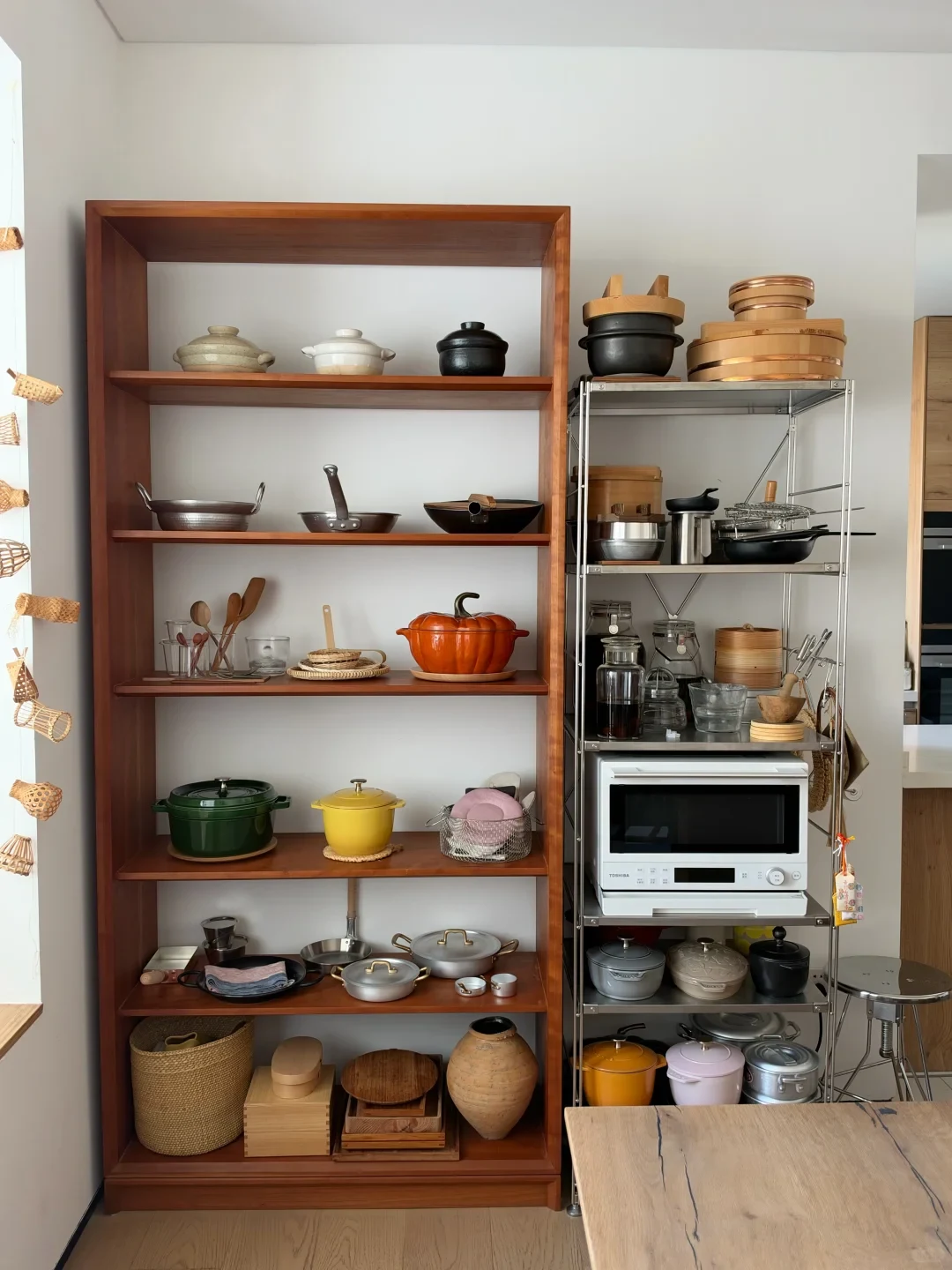 A kitchen corner featuring a tall wooden open shelf and a stainless steel shelving unit filled with organized cookware and kitchenware, including cast-iron Dutch ovens, pans, ceramic pots, glassware, a pumpkin-shaped cocotte, and a white microwave oven.