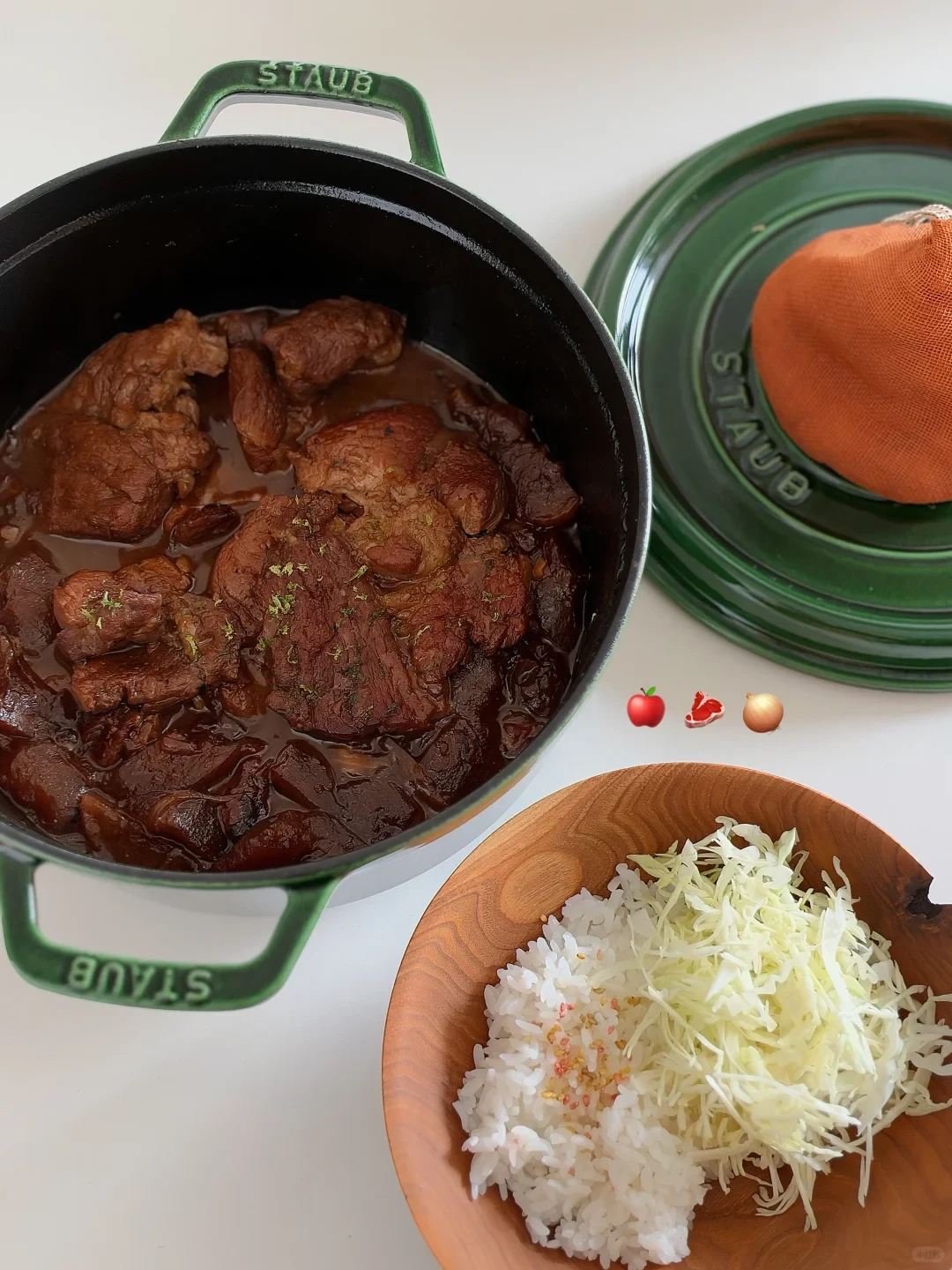 A top-down view of braised pork cooked in a green Staub cocotte, served alongside a wooden bowl containing white rice and shredded cabbage, with the pot lid resting next to it.