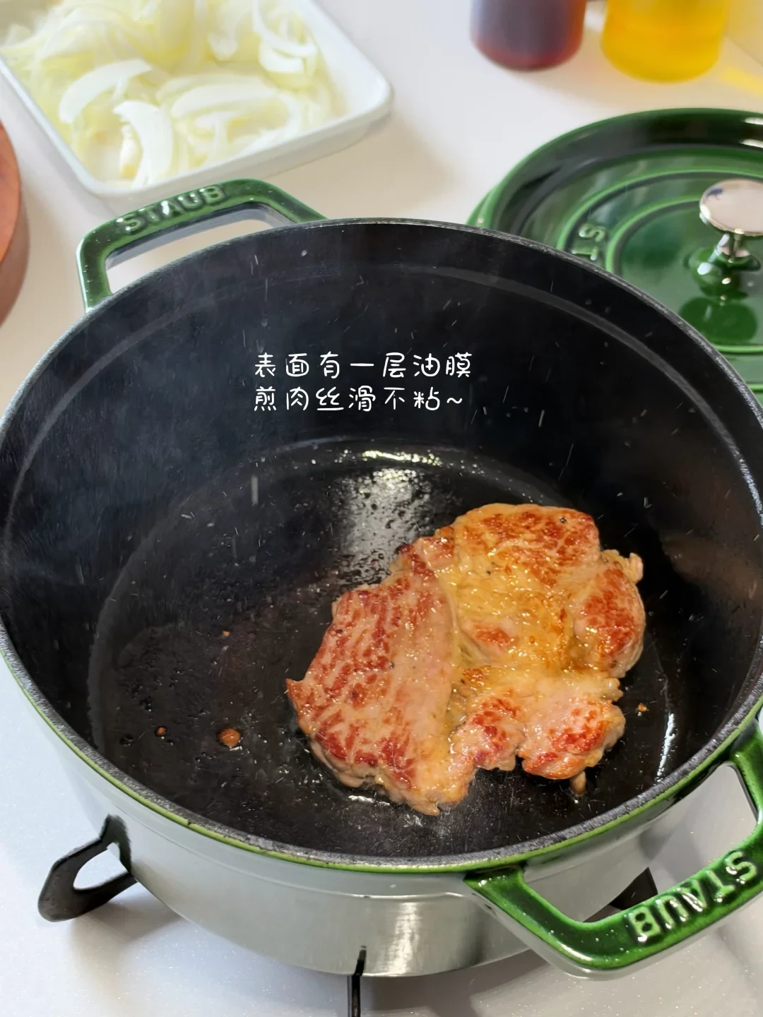A piece of meat searing to a golden brown inside a green Staub cast iron pot on a stovetop.