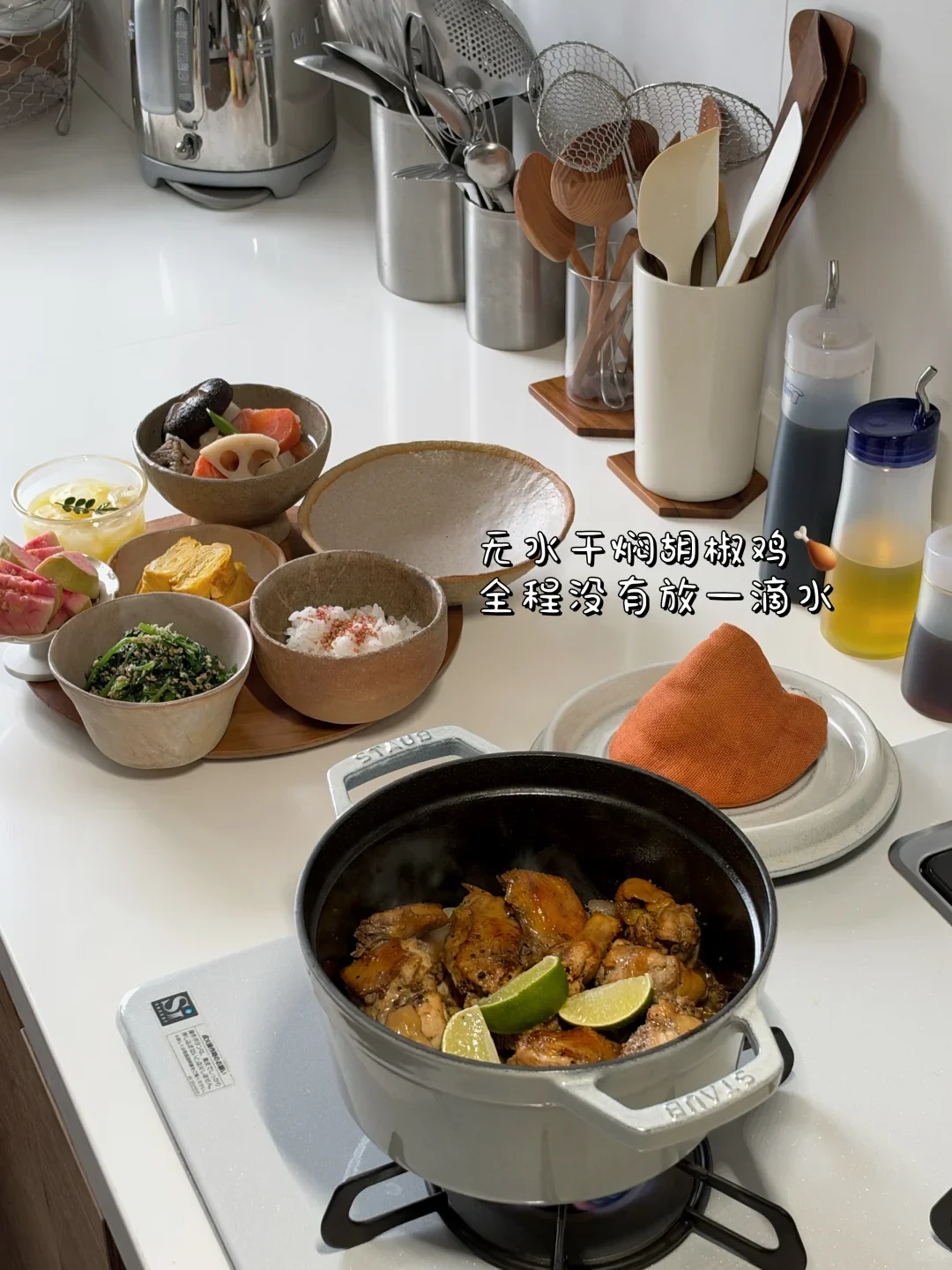 A kitchen counter spread featuring a Staub cast-iron cocotte of braised pepper chicken garnished with lime wedges on a burner, alongside several small bowls containing rice, tamagoyaki, vegetables, and fruit arranged on a wooden tray.
