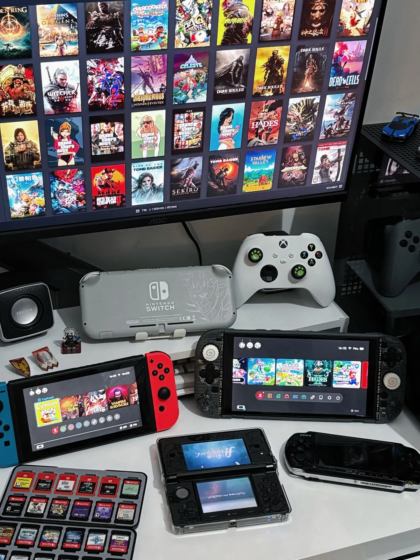 A gaming desk setup featuring a large PC monitor displaying a digital game library above an array of handheld consoles. On the desk are a white Xbox controller, a special edition grey Nintendo Switch Lite, a standard Nintendo Switch with red and blue Joy-Cons, a handheld console running a Switch interface, an open Nintendo 3DS displaying Fire Emblem Fates, a black Sony PSP, and an organizer tray filled with Nintendo Switch game cartridges.