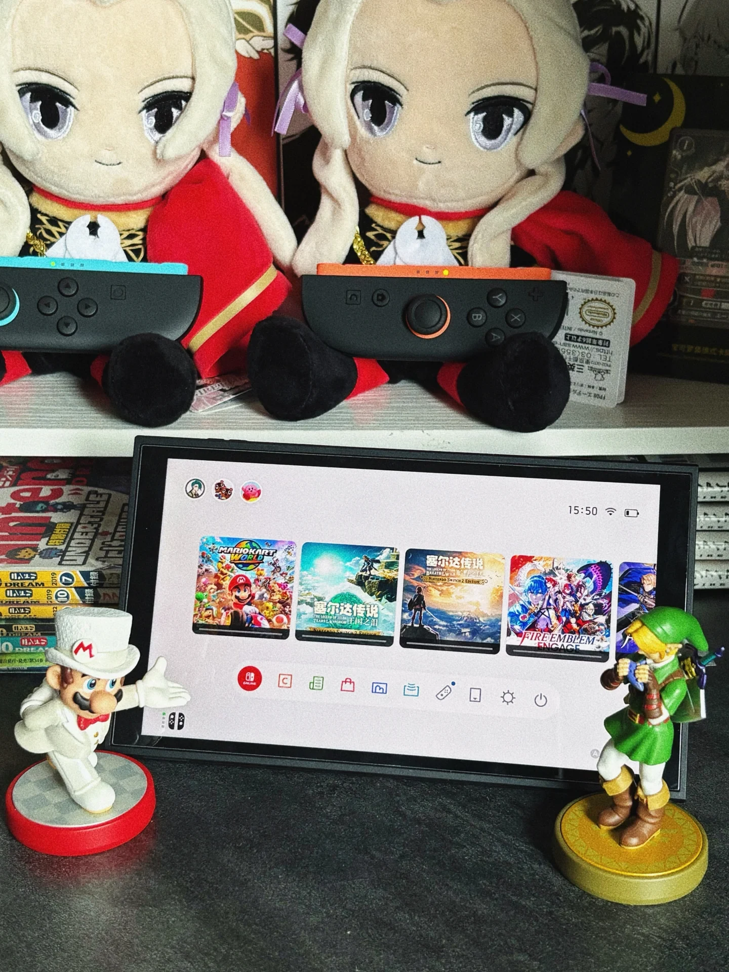 A Nintendo Switch console sitting upright on a desk, flanked by a Wedding Mario amiibo on the left and a Link amiibo on the right. On a shelf above, two Edelgard plushies sit holding Joy-Con controllers.