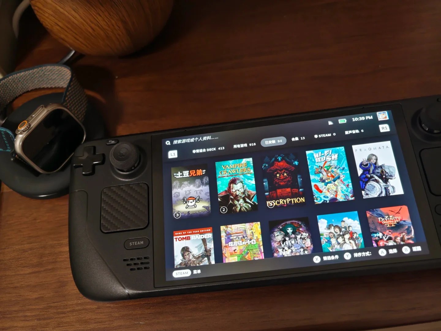 A Steam Deck handheld gaming console resting on a wooden desk displaying its game library interface, positioned beside an Apple Watch Ultra on a charging stand.