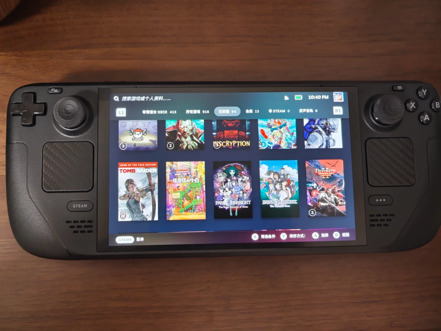 A black Steam Deck handheld gaming console sits on a wooden surface, displaying its game library interface with title covers including Inscryption, Tomb Raider, and Disco Elysium.