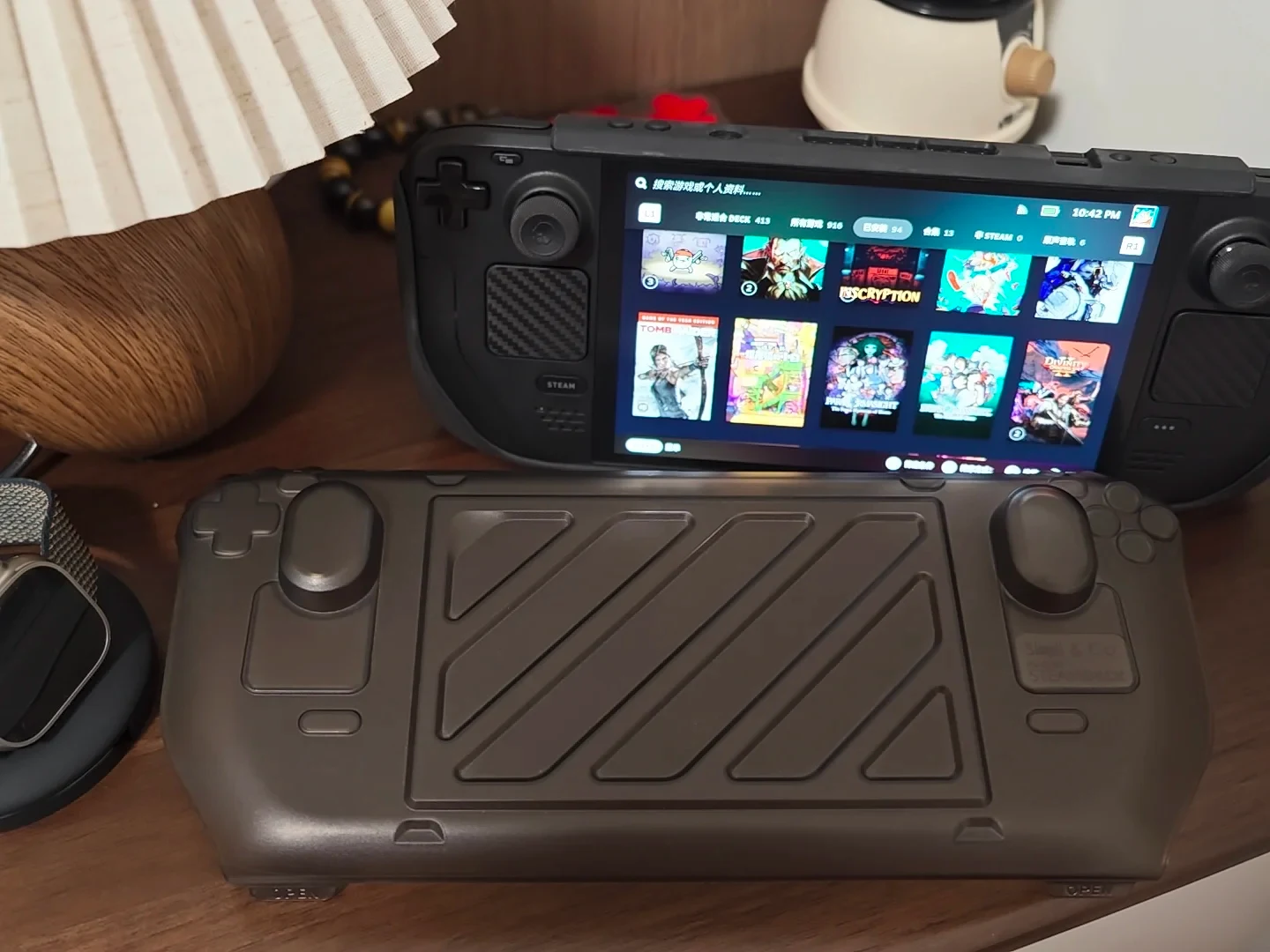 A Steam Deck handheld console sits on a wooden desk with a black matte protective hard front cover resting in front of it.