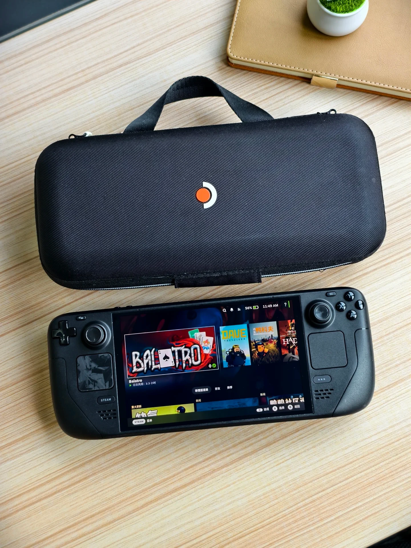 A black Steam Deck handheld gaming console displayed on a light wood desk next to its zippered carrying case, showing the game Balatro on the screen.