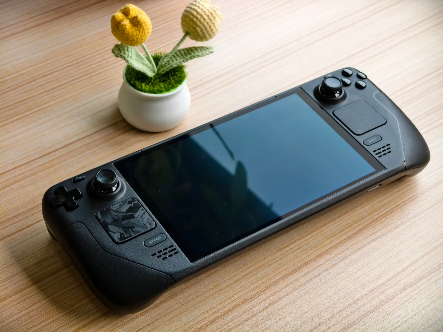 A black Steam Deck handheld gaming console resting on a light wood-grain table next to a small white pot with crocheted yellow flowers.