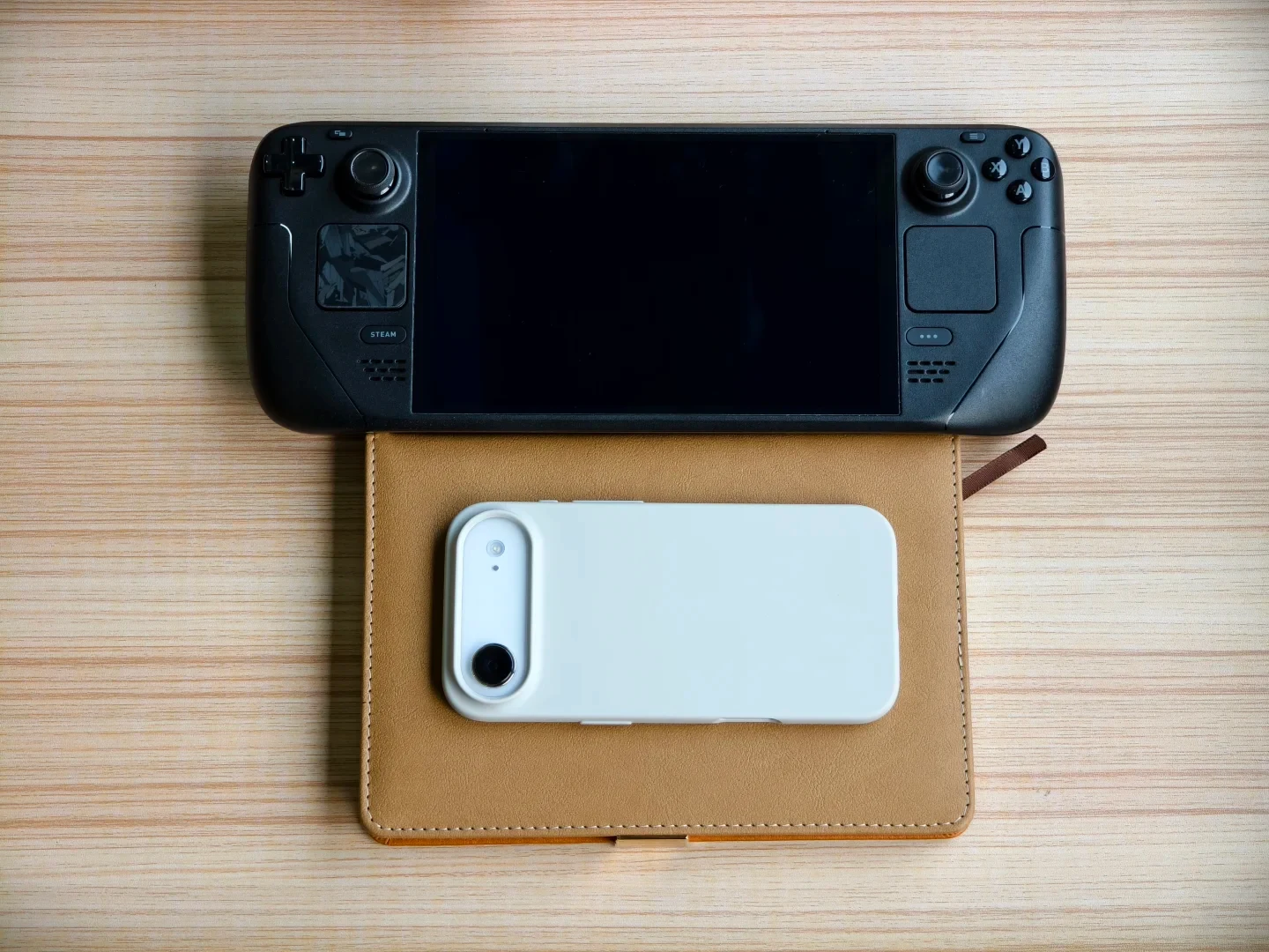 A black Steam Deck handheld console positioned horizontally above a light brown notebook, on top of which rests a horizontal white smartphone.