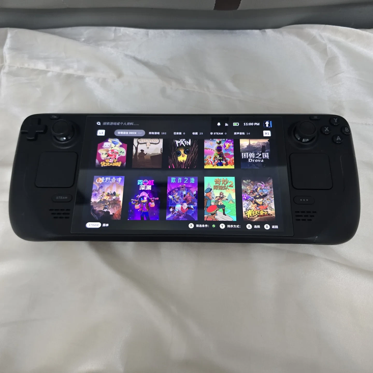 A black Steam Deck handheld gaming console resting on a white fabric surface with its screen turned on, displaying the SteamOS game library with several game titles visible.