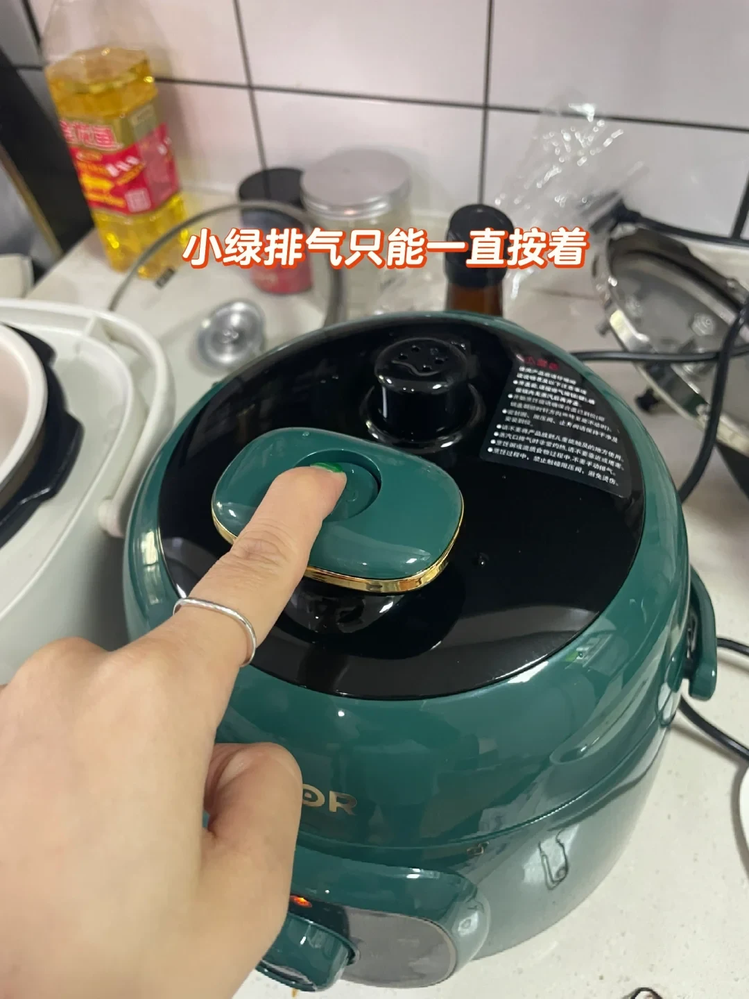 A person's finger presses down on the steam release button on the lid of a dark green electric pressure cooker sitting on a kitchen counter.