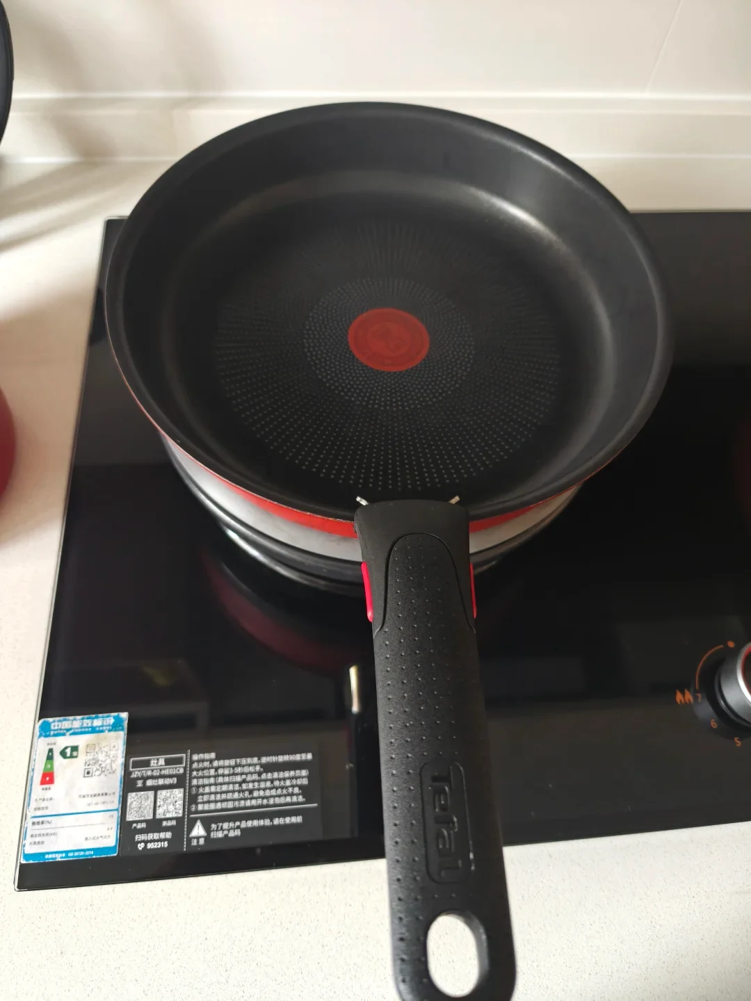 A black non-stick Tefal frying pan with a red exterior rim and detachable handle placed on a heat diffuser plate over a black stovetop.