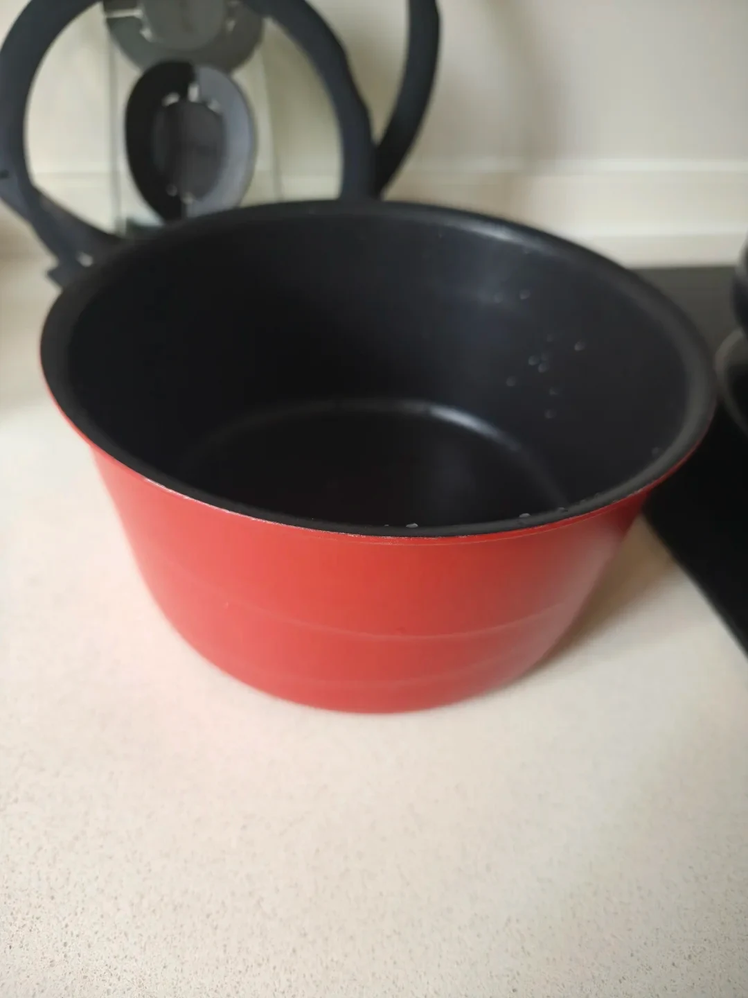 A round red Tefal saucepan without a handle sitting on a white countertop with a glass and silicone lid in the blurred background.