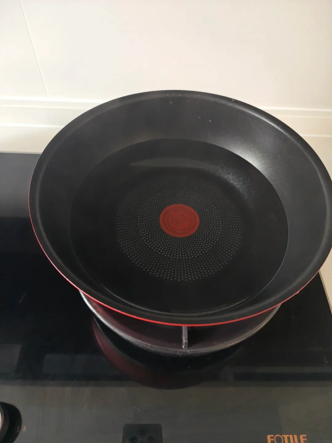 A round Tefal nonstick frying pan filled with water resting on a heat diffuser disc on a black gas cooktop without its detachable handle.
