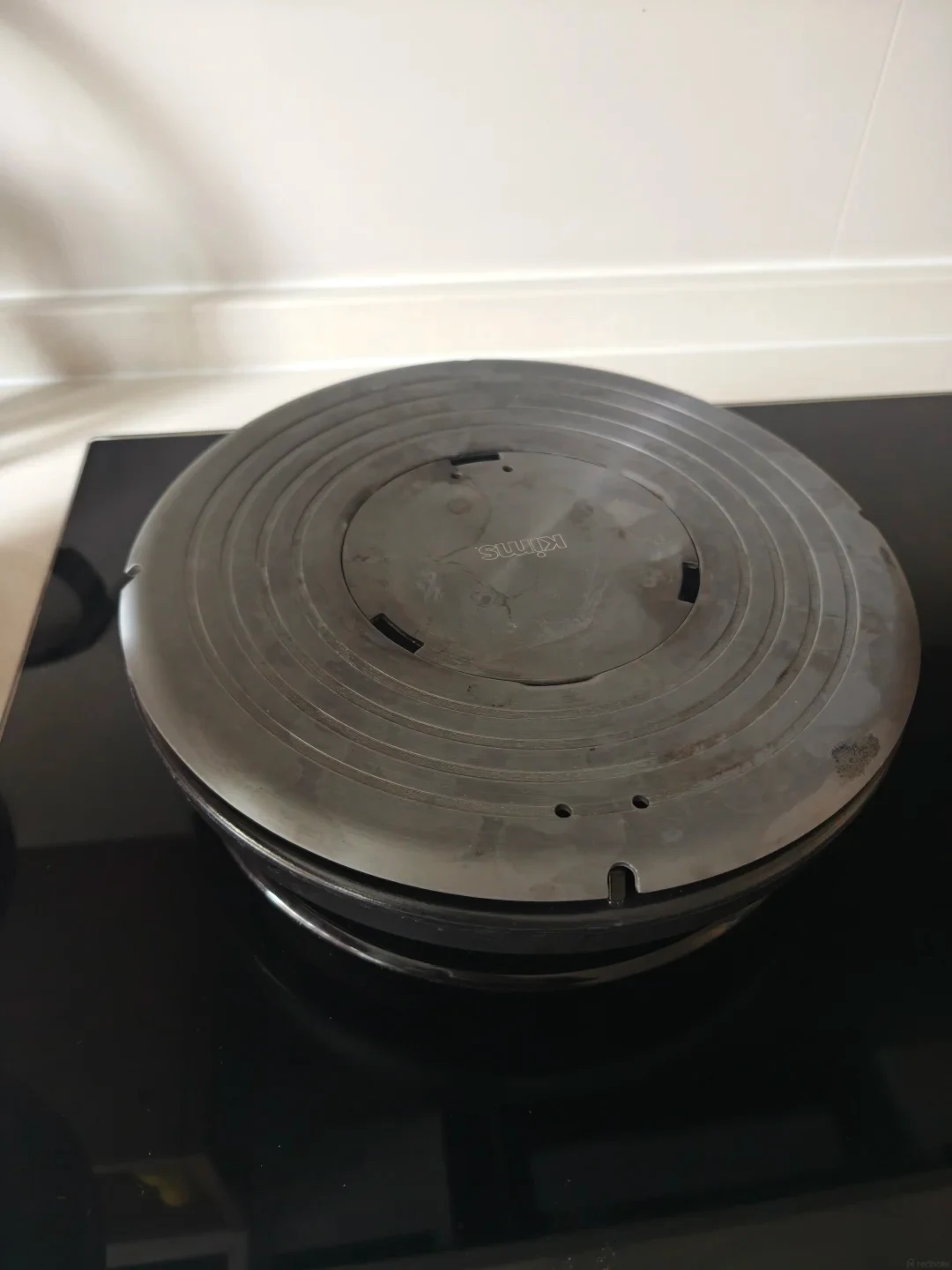 A round, dark metal heat diffuser plate with concentric grooves placed on a stovetop burner.