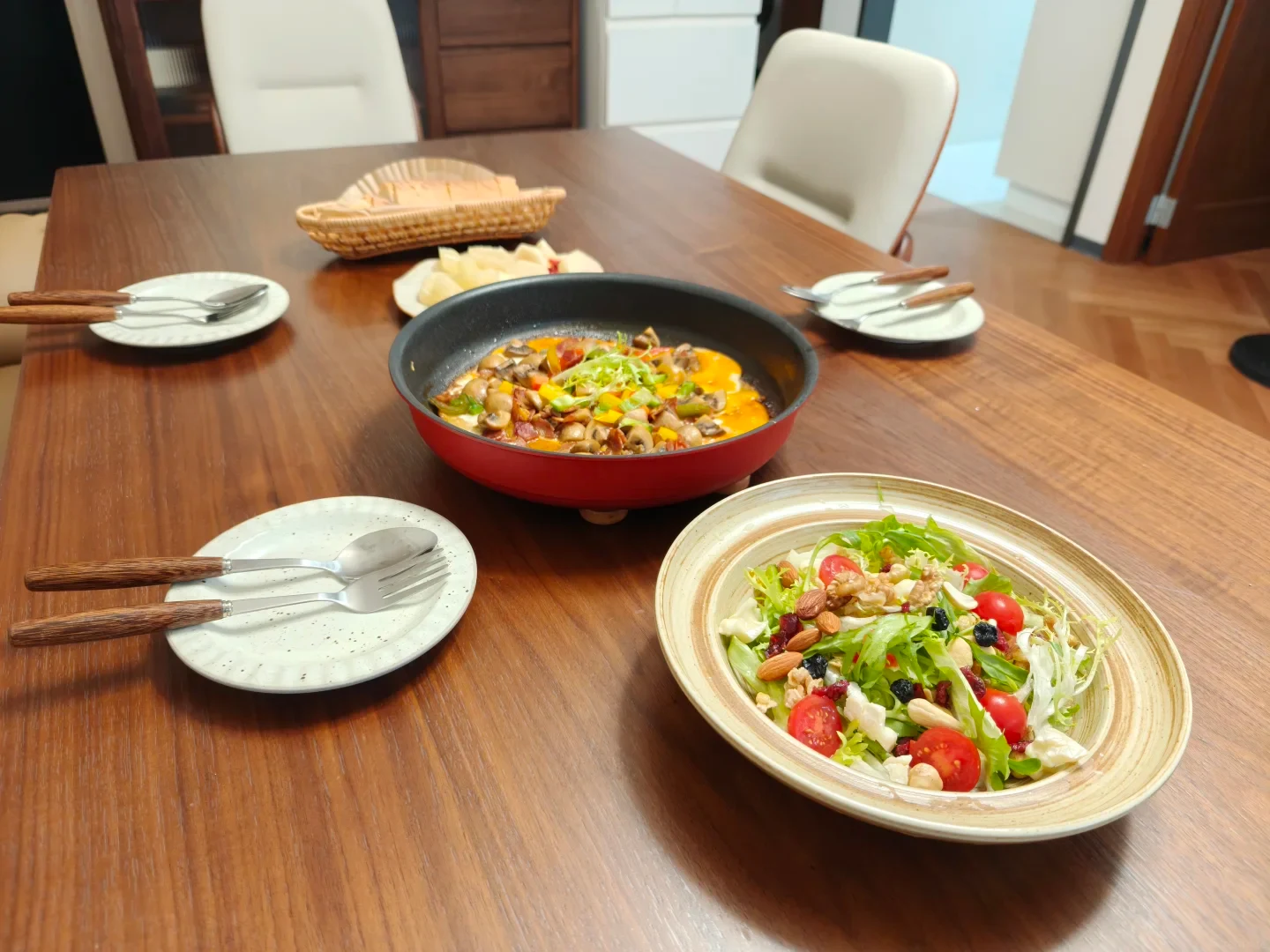 Red Tefal non-stick pan filled with cooked food and served directly on a wooden dining table with its handle removed, beside a bowl of salad and place settings.