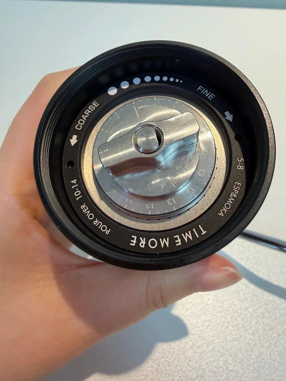 A hand holds a black Timemore coffee grinder adjustment dial with a silver center and numbered grind-setting ring. The dial is marked COARSE and FINE, with arrows showing adjustment directions.