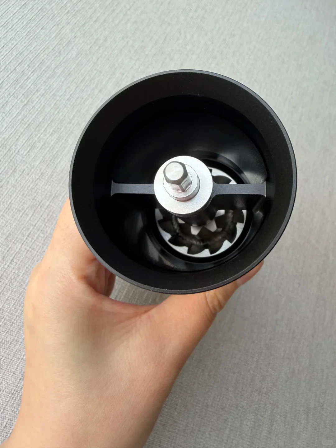 A hand holds the open grinder, showing its central metal shaft, crossbar and burr assembly inside the black hopper.