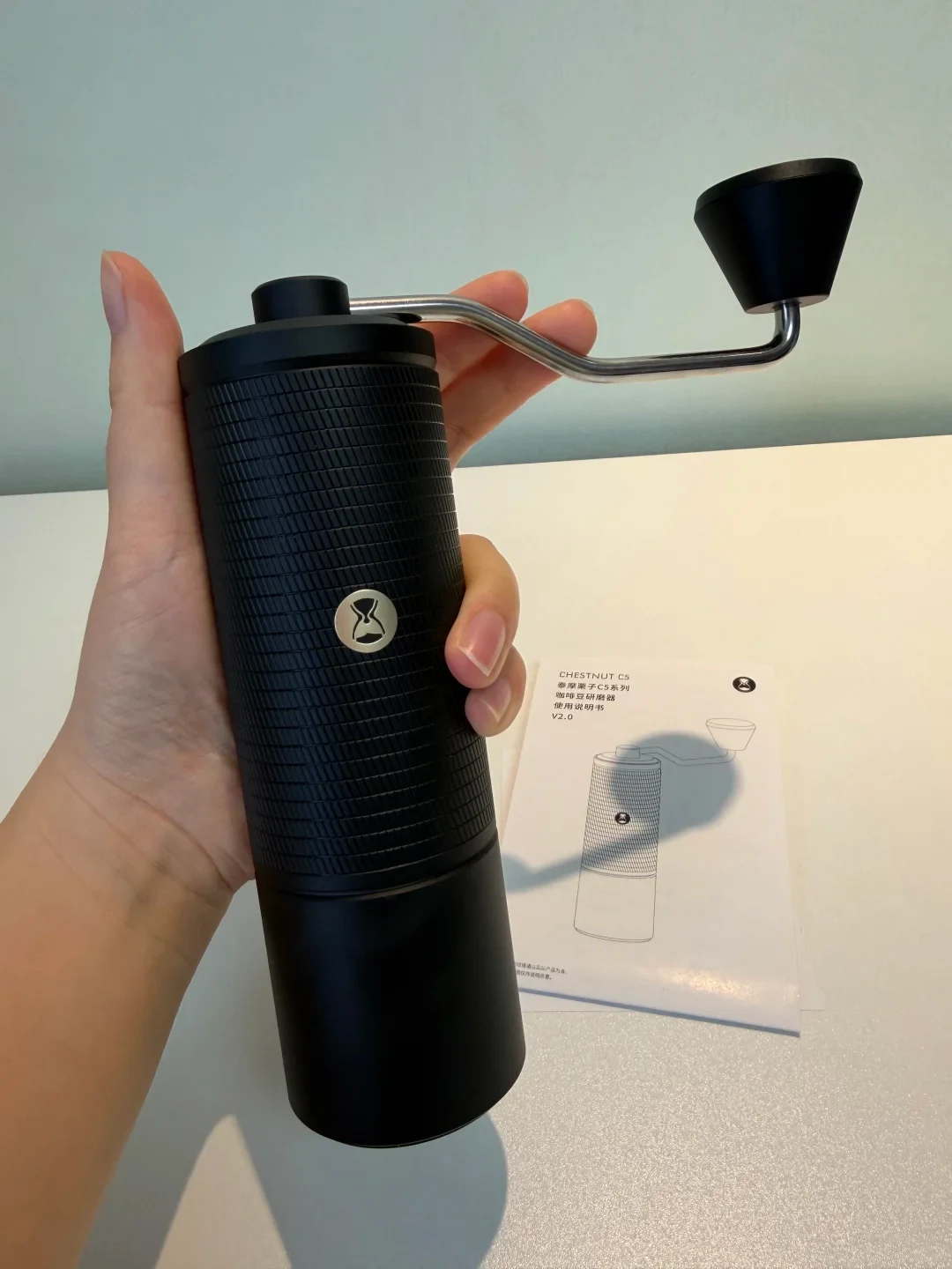 A hand holds a black, ridged manual coffee grinder with a metal crank and black knob above a tabletop. Its instruction booklet, showing a line drawing of the grinder, lies behind it.