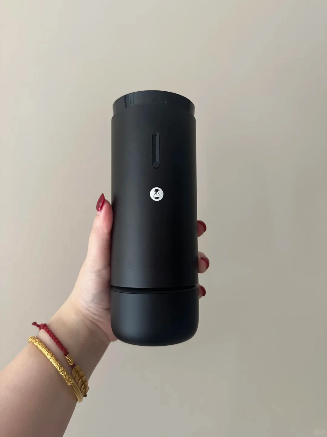 A hand with red-painted nails and bracelets holds a matte black cylindrical device with a narrow vertical indicator and a small white hourglass-like logo against a plain wall.