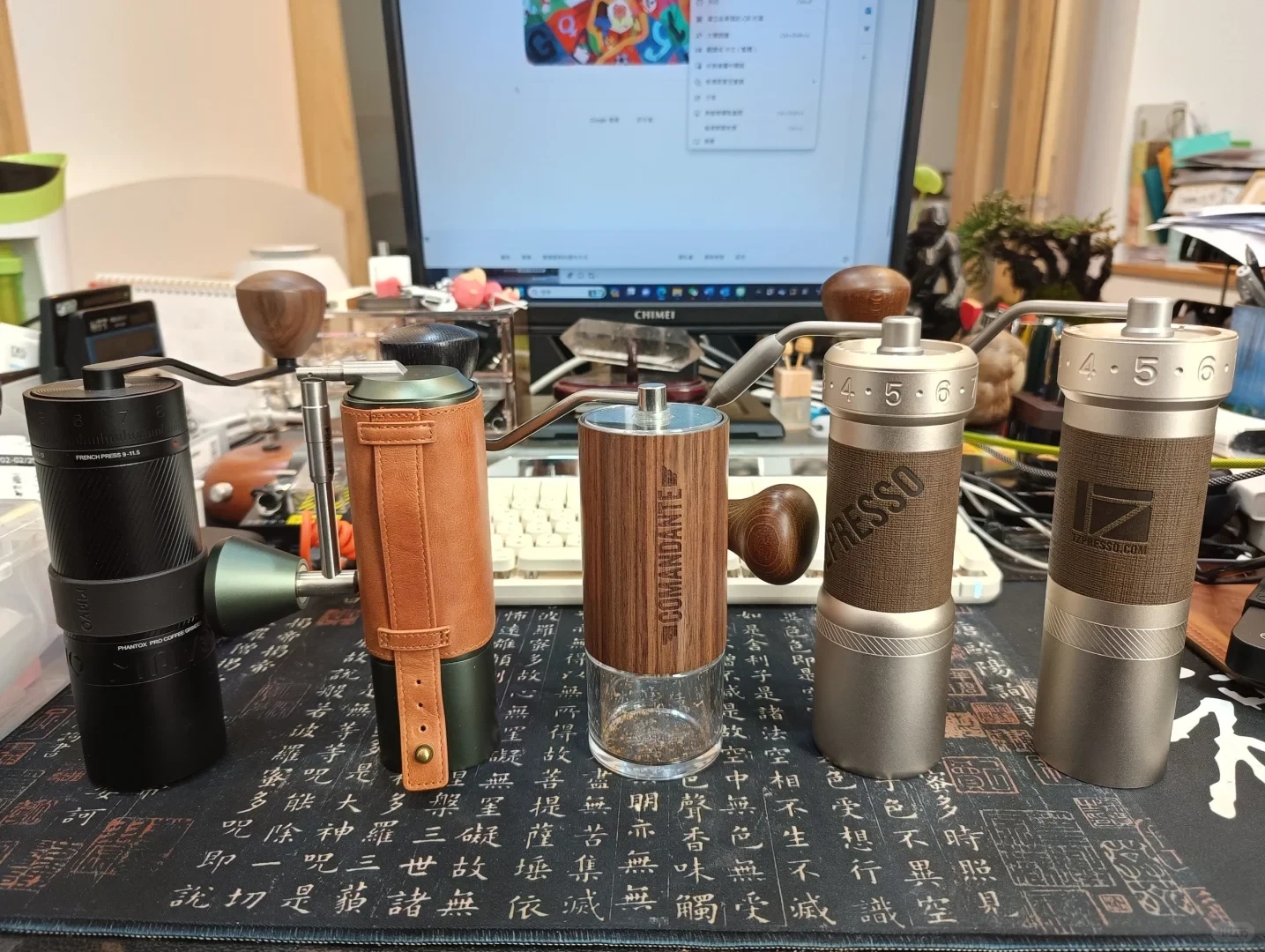 Five manual coffee grinders in different colors and styles are lined up on a desk, with a computer, keyboard, and other items behind them.