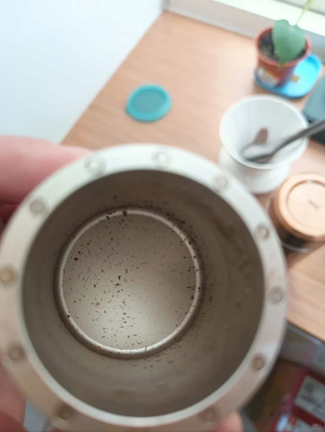 A hand holds a silver-colored grinder catch cup with scattered coffee grounds on its inner wall and bottom.