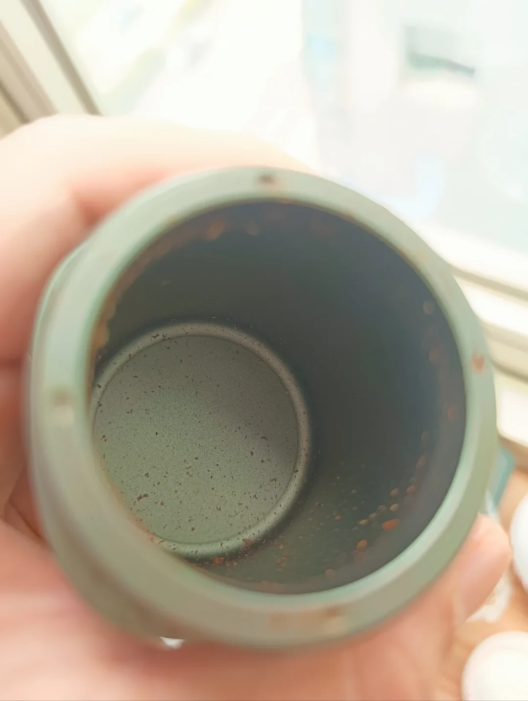 A hand holds a green grinder catch cup with scattered coffee grounds and small clumps on its inner wall and bottom.
