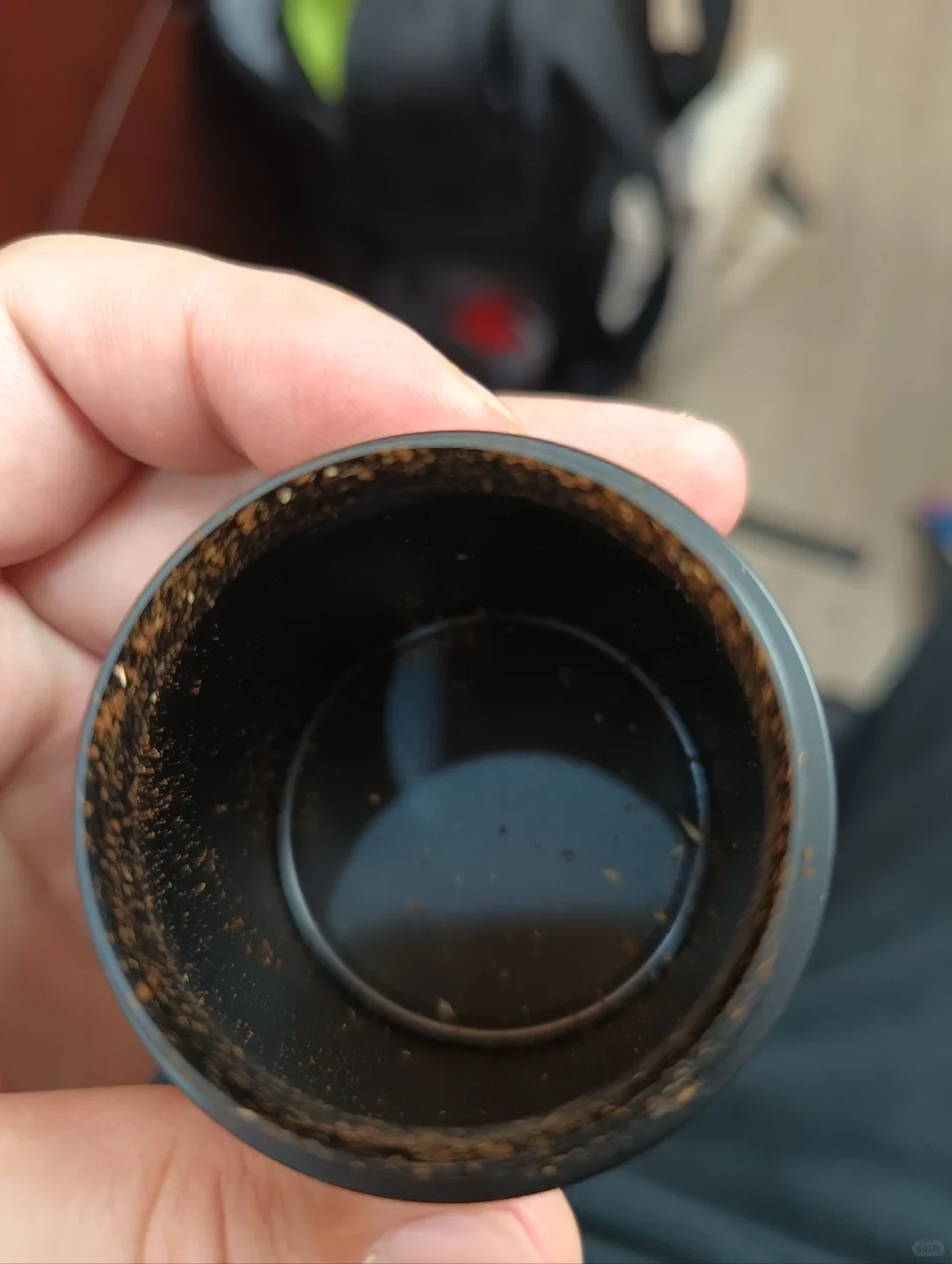 A hand holds a black grinder catch cup with coffee grounds accumulated around the upper inner wall and rim.