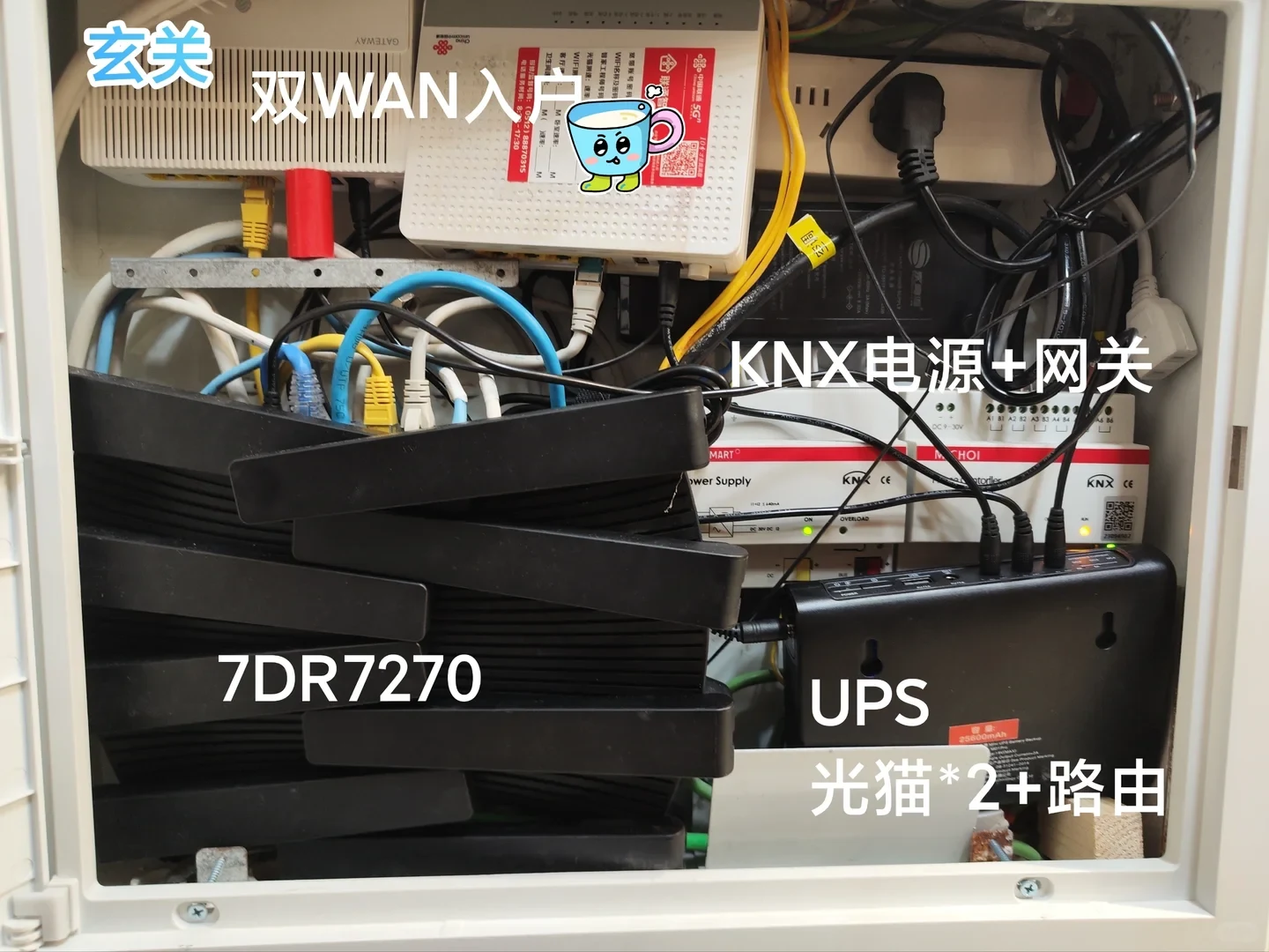 Open entryway communications cabinet containing two optical network modems at the top, a black 7DR7270 router, KNX power supply and gateway hardware, a UPS, and dense Ethernet and power cabling.