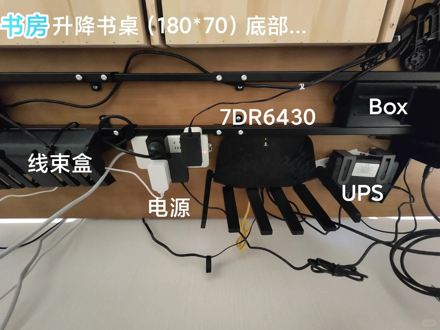 Underside view of a wooden standing desk showing mounted network equipment, including a wire management box, a power strip with adapters, an inverted black multi-antenna 7DR6430 router, and a compact UPS unit.
