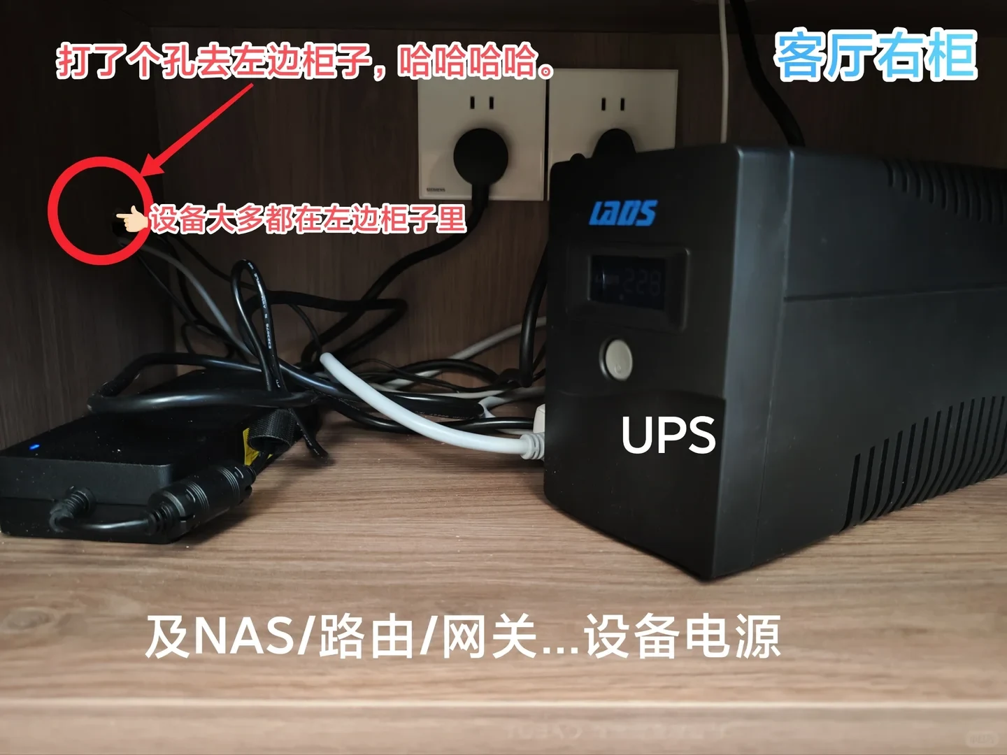 A black UPS unit and power bricks inside a wooden TV cabinet shelf, with power cords passing through a drilled hole on the left wall.