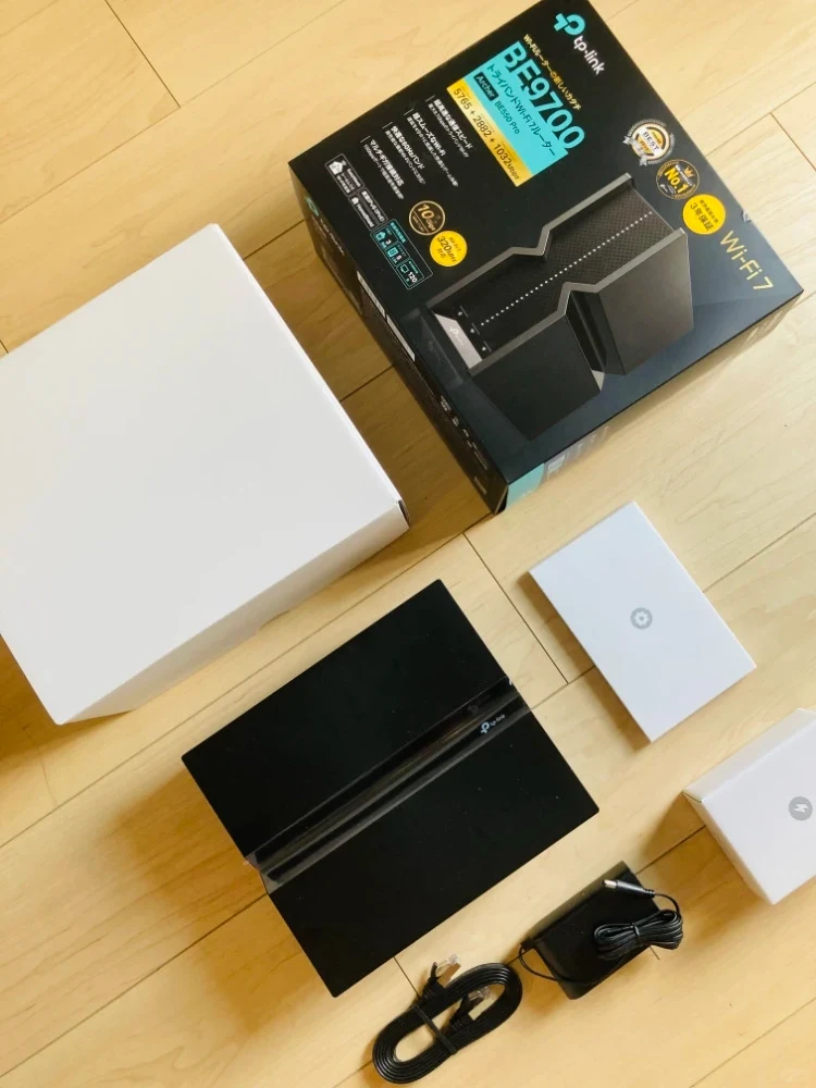 Unboxing display of a TP-Link Archer BE550 Wi-Fi 7 router on a wooden floor, showing the retail packaging box, inner boxes, the black router unit, an Ethernet cable, and the power adapter.