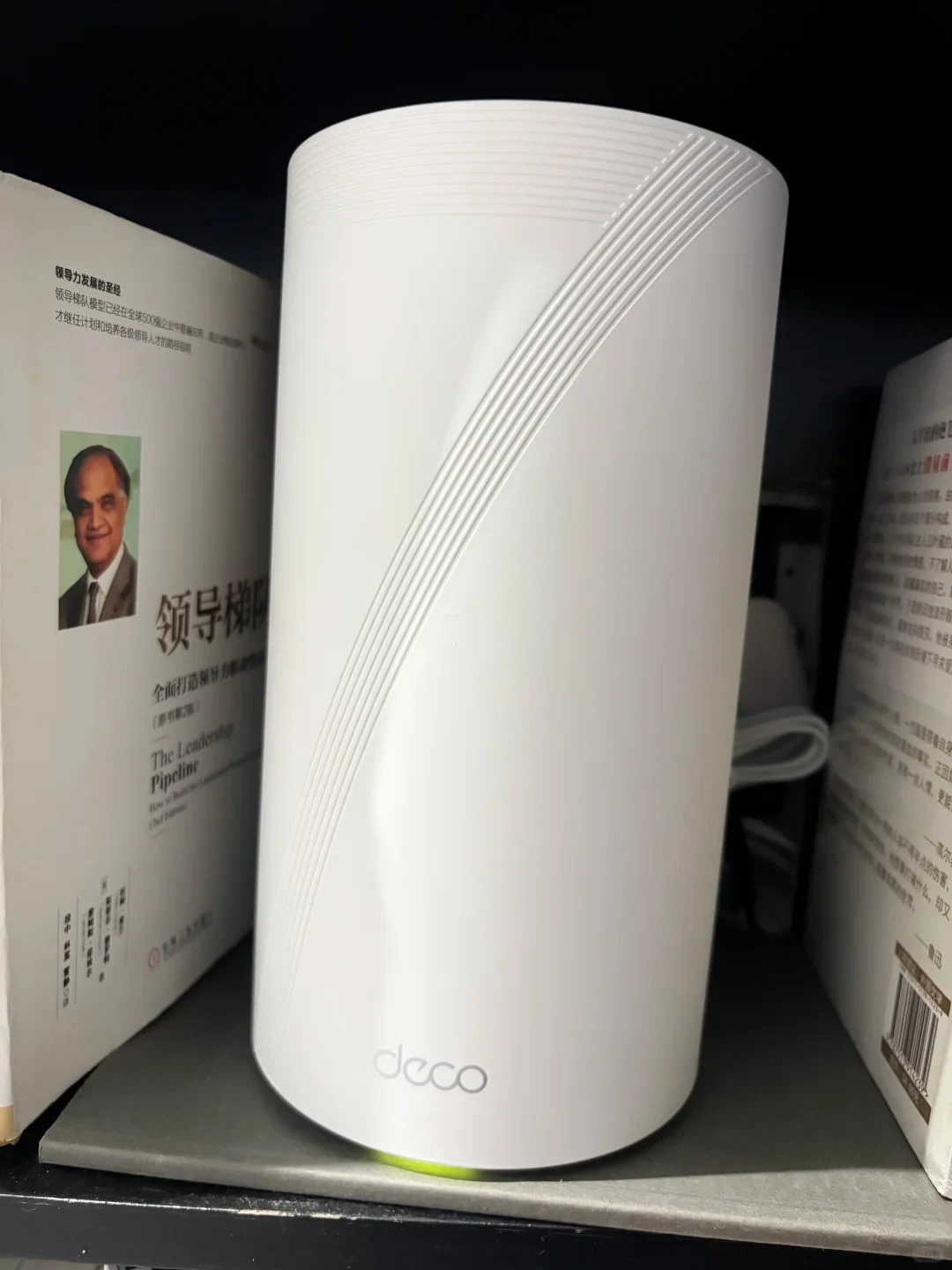 A white TP-Link Deco BE95 cylindrical mesh router placed on a bookshelf between books, glowing yellow-amber at the base indicator light.