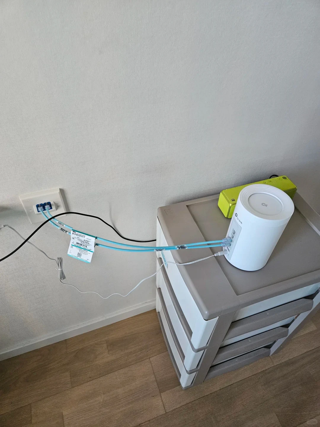 A white cylindrical TP-Link Deco router placed on a plastic storage drawer unit, connected via two light blue Ethernet cables to a dual-port wall network plate.