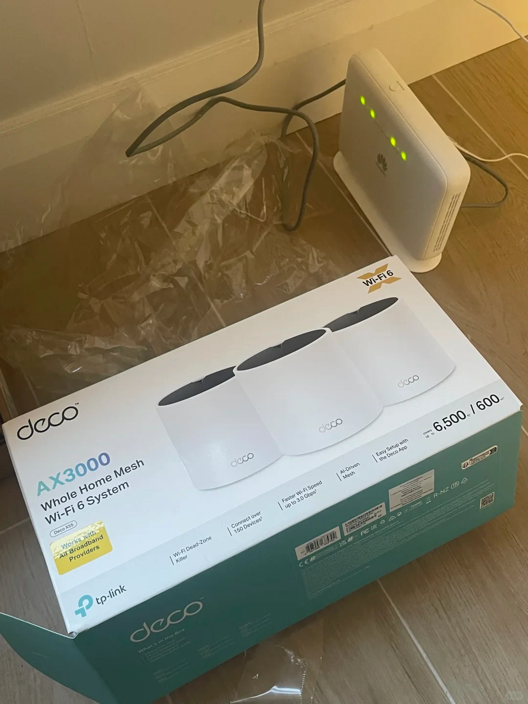 A TP-Link Deco AX3000 Whole Home Mesh Wi-Fi 6 System box resting on a light wood floor next to an upright white Huawei router with green indicator lights illuminated.
