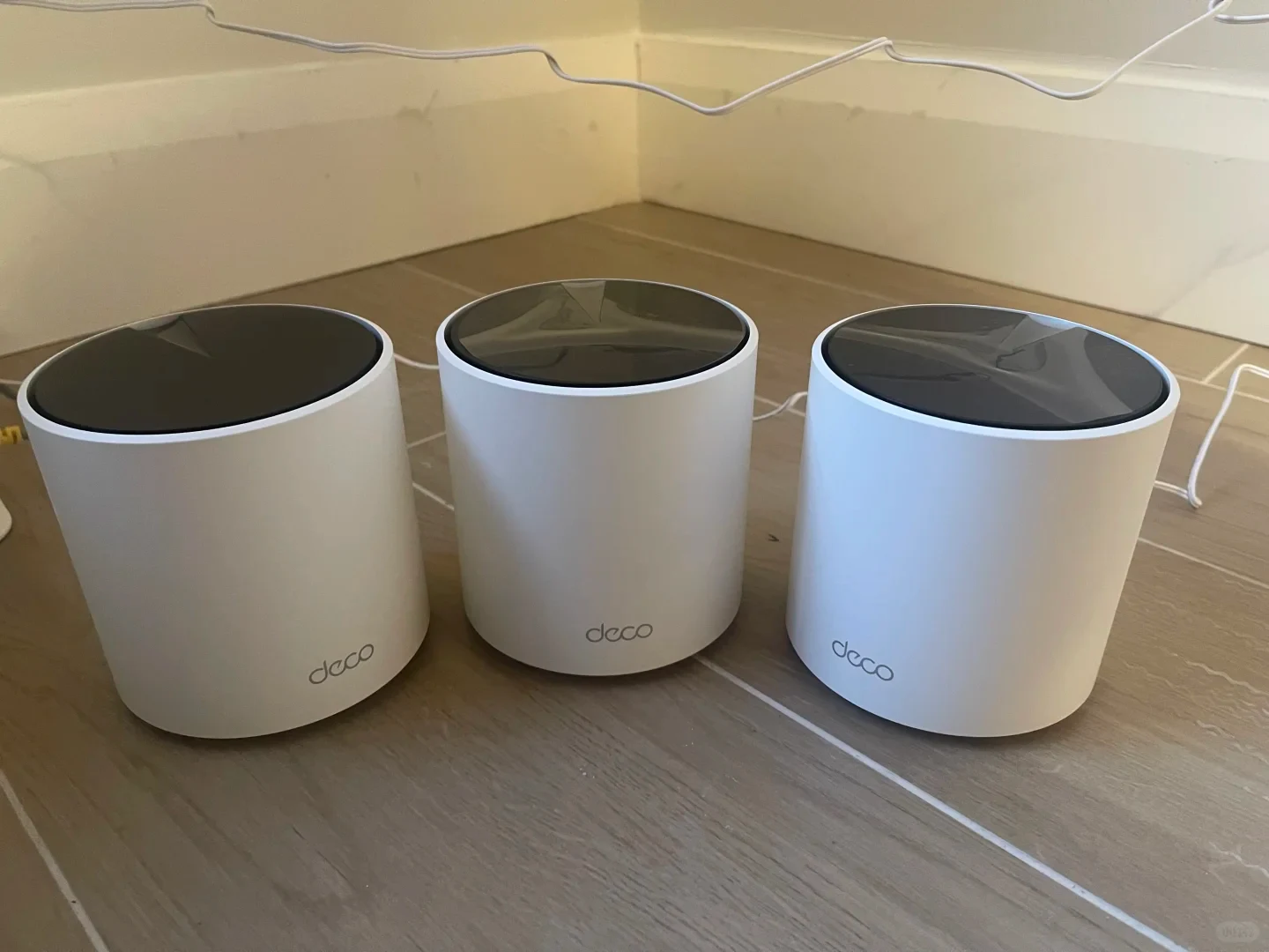 Three white cylindrical Deco mesh Wi-Fi units sitting side by side on a tiled floor with white cables visible behind them.