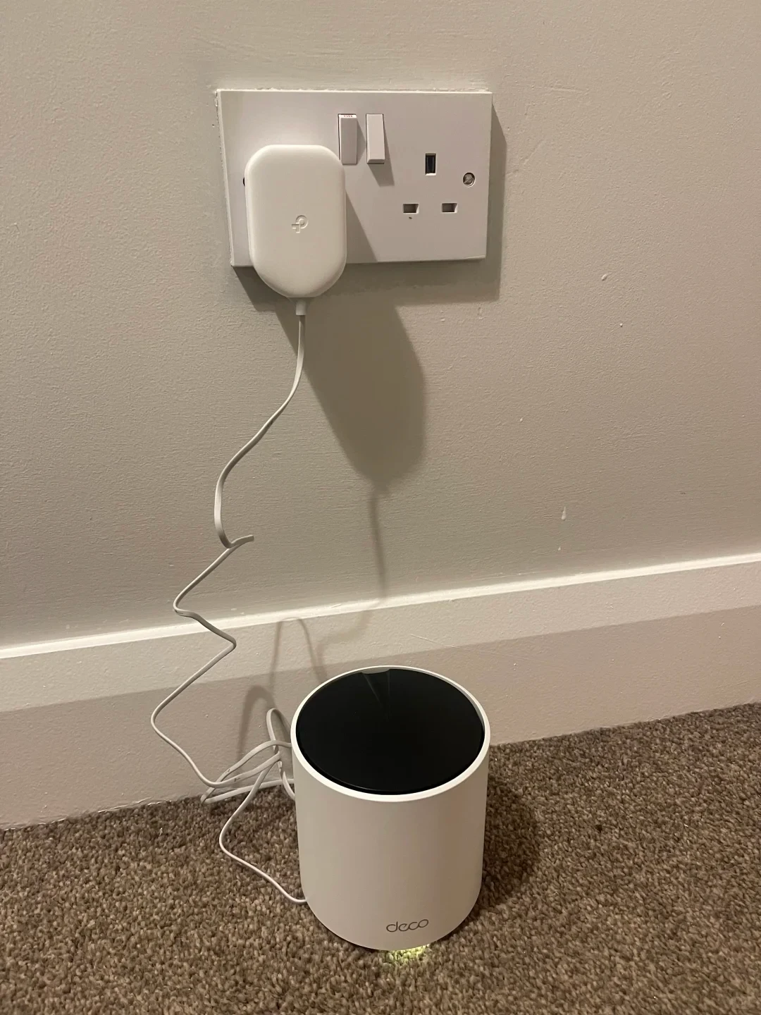 A white cylindrical TP-Link Deco mesh Wi-Fi router sitting on a carpeted floor next to a white baseboard, connected by a power cable to an adapter plugged into a dual UK wall outlet.