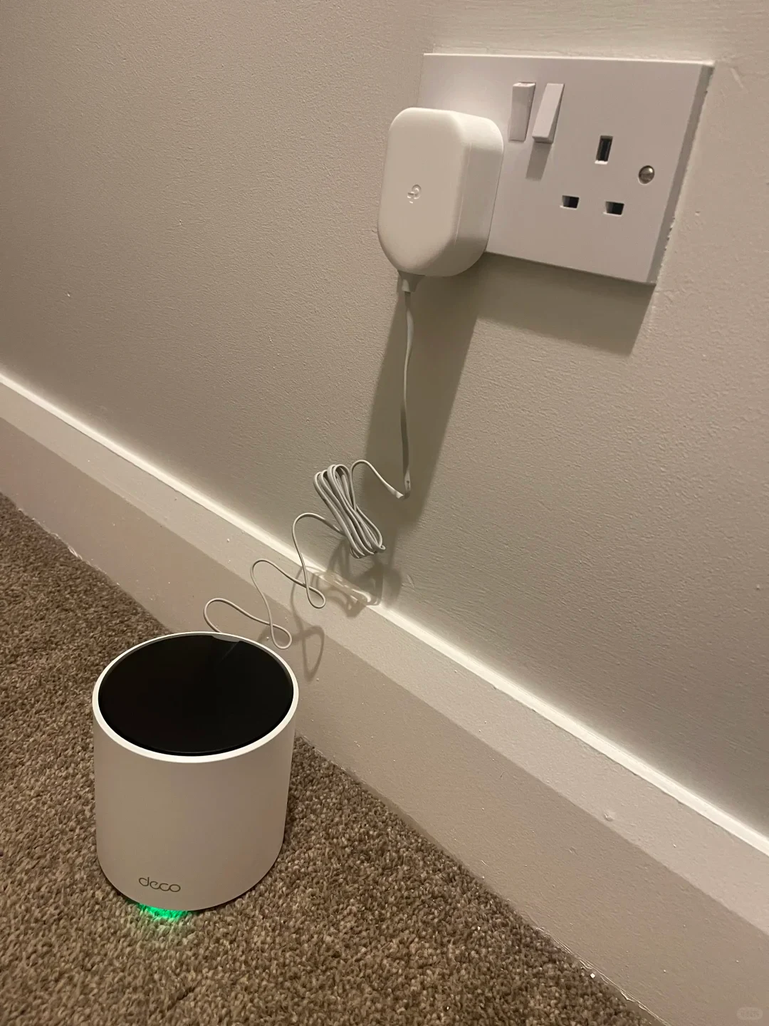 A white cylindrical TP-Link Deco mesh Wi-Fi router sitting on a carpeted floor with a green indicator light glowing underneath. A thin white power cord connects the router to a white power adapter plugged into a dual UK wall socket mounted on a white wall above a baseboard.